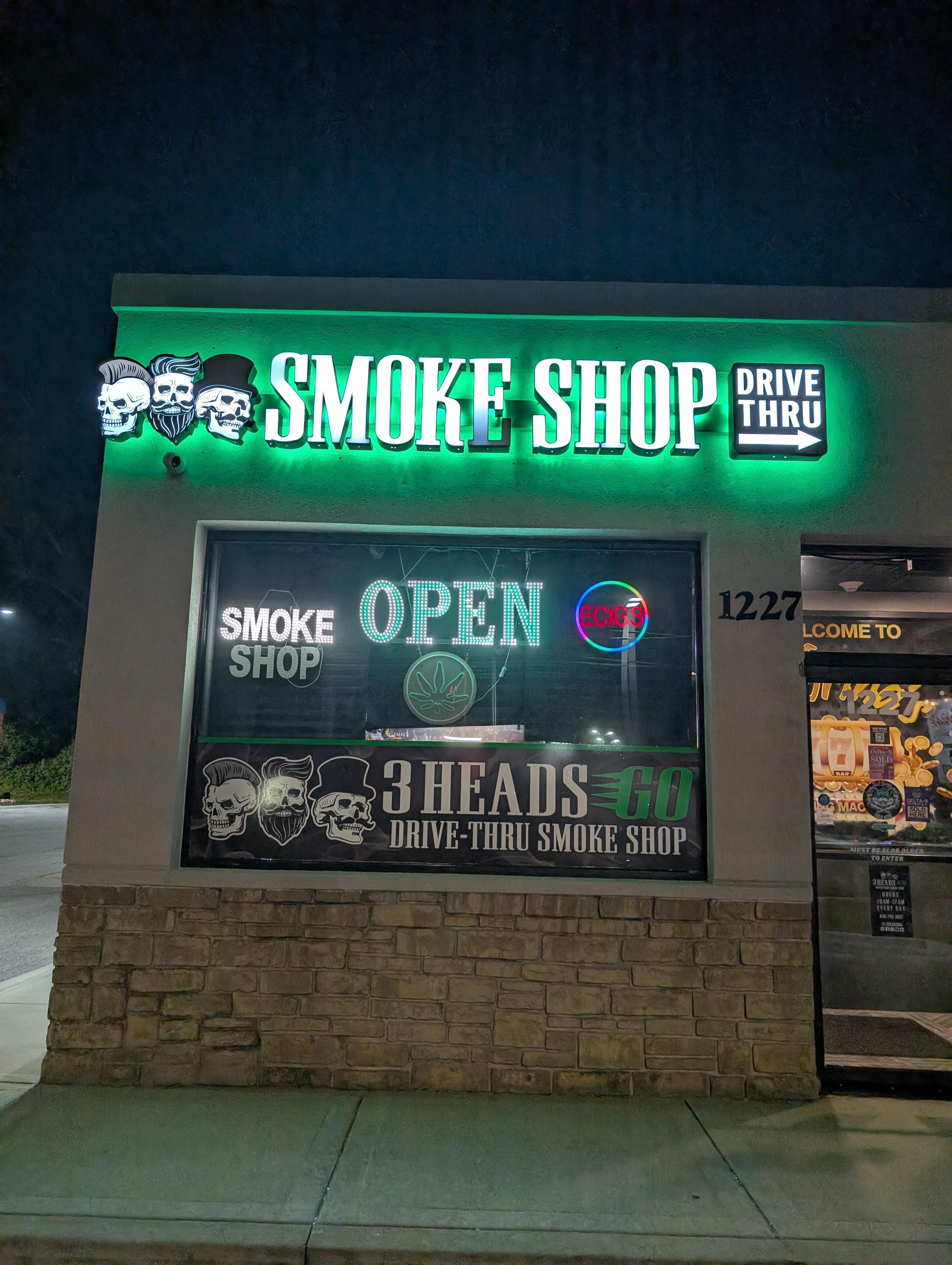 The 3 Heads Go storefront at 1227 Glen Ellyn Rd in Glendale Heights at night — the green-lit three-skull SMOKE SHOP sign with its DRIVE THRU arrow above the window