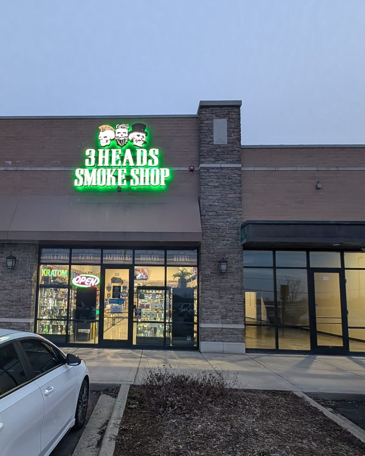 The green neon 3 Heads Smoke Shop sign with its three skulls lit at dusk above the Elgin storefront