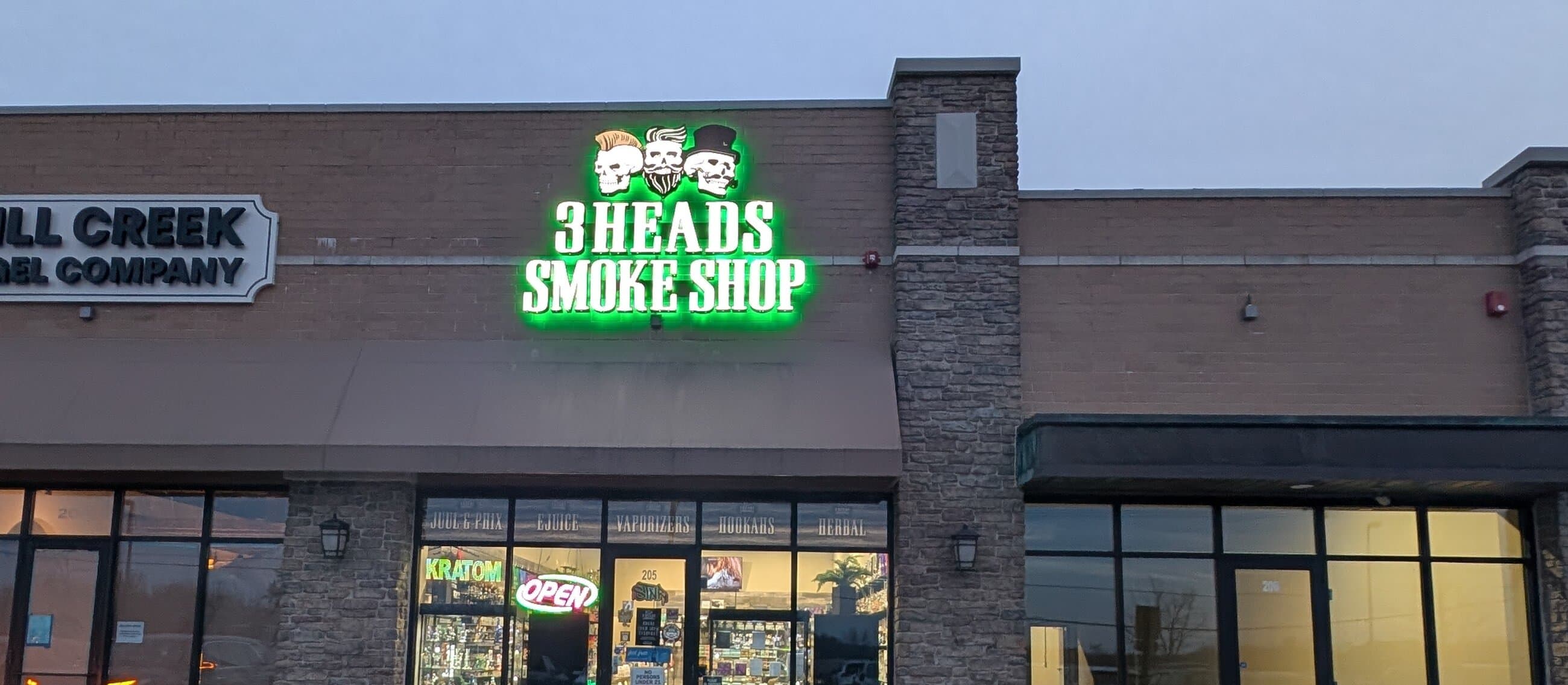 The 3 Heads Smoke Shop flagship in Elgin at dusk, with the green neon three-skull sign lit above the entrance